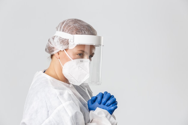 Profile of hopeful middle-aged female doctor in personal protective equipment, face shield, gloves and respirator, praying or pleading.