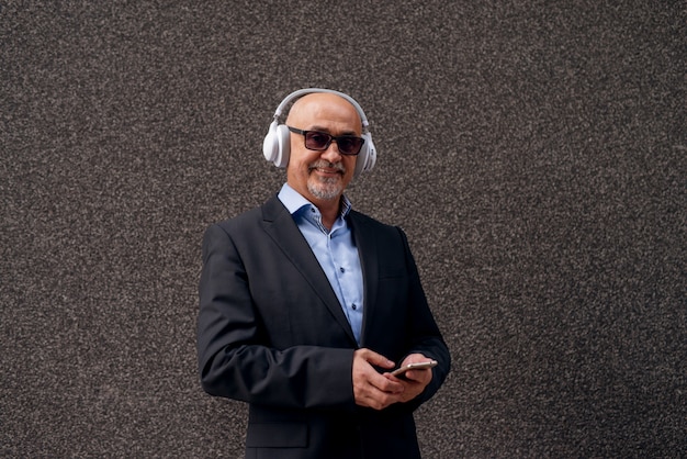 成熟した陽気なプロのエレガントなビジネスマンのプロファイルは、ヘッドフォンで電話で音楽を聴いています。