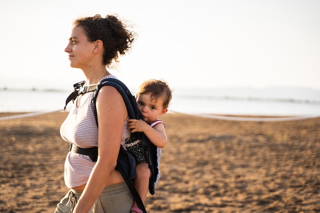 Profilo di una madre che cammina portando un bambino sulla schiena in un marsupio sulla spiaggia