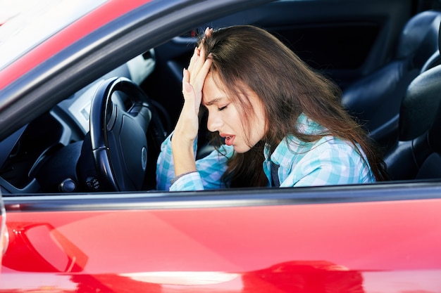 赤い車を運転している神経質な女性のプロフィール、運転中のストレス。目を閉じて手に寄りかかって緊張した女性、交通渋滞。車内のブルネットの女性の頭と肩