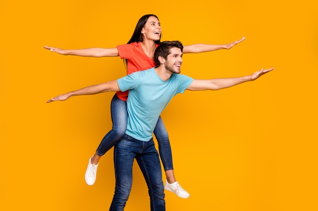 Foto del profilo del ragazzo divertente portare lady piggyback trascorrere l'estate insieme diffondere le mani come ali indossare casual elegante blu arancione t-shirt jeans isolato giallo parete di colore