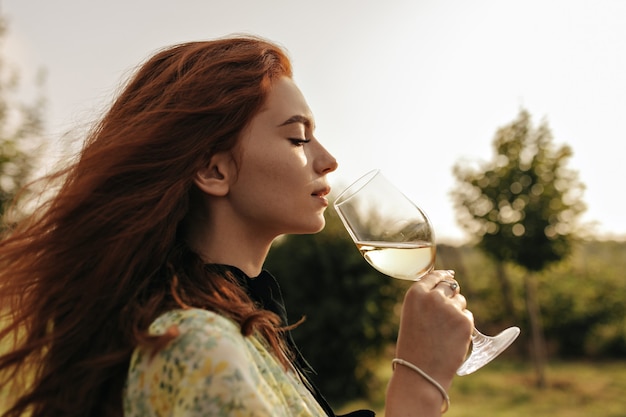 Foto del profilo di una giovane donna dai capelli rossi in eleganti abiti verdi e braccialetto che tiene in mano un bicchiere di champagne e beve all'aperto