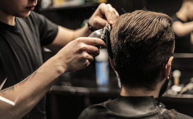 Profile photo of a young barber trimming his customerÃ¢ÂÂs hair with an electric shaver and a comb in a barbershop.