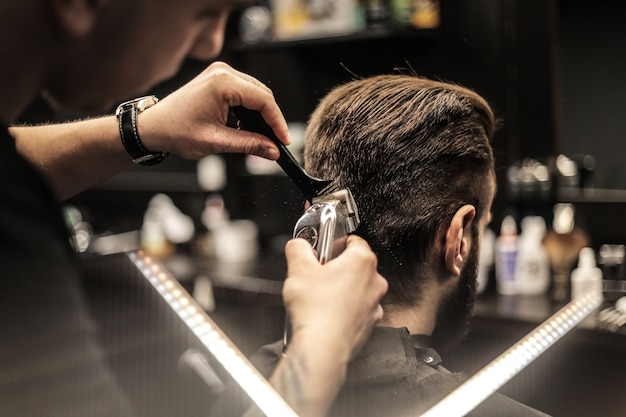 Profile photo of a young barber trimming his customerÃ¢ÂÂs hair with an electric shaver and a comb in a barbershop.