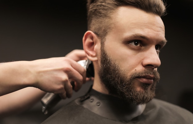 Profile photo of a young barber trimming his customerÃ¢ÂÂs hair with an electric shaver and a comb in a barbershop.