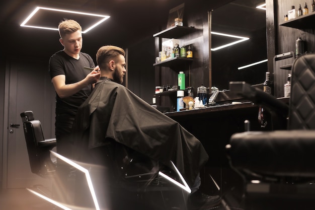 Profile photo of a young barber trimming his customerã¢ââs hair with an electric shaver and a comb in a barbershop.