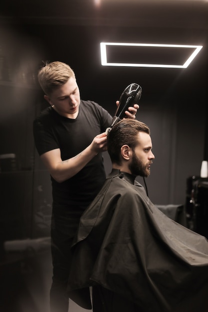 Photo profile photo of a young barber trimming his customerã¢ââs hair with an electric shaver and a comb in a barbershop.