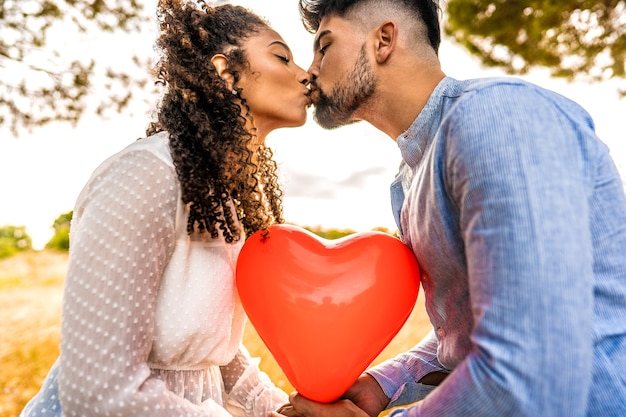 写真 彼らの間で赤いハート型の風船に太陽のバックライト効果で自然の中で日没にキスする愛の多民族カップルのプロフィール写真。 2人の異性愛者の若者の夕暮れ時のロマンチックなシーン