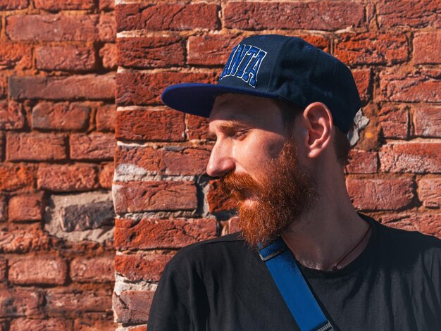 Photo profile portrait of bearded millennial hipster man in snapback on brick wall urban background stylish confident authentic guy with red beard casual clothes black tshirt walking on sunny city street