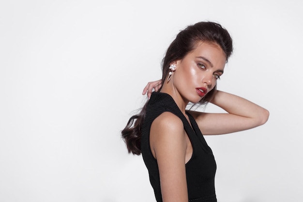 Profile portrait of a beautiful brunette woman with tanned skin dressed in black dress, with makeup, wears luxurious jewelry, looking at camera, on a white background.