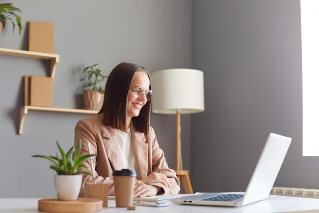 Profile portrait of charming adorable woman with brown hair wearing beige jacket working on laptop looking at notebook display with toothy smile having online meeting