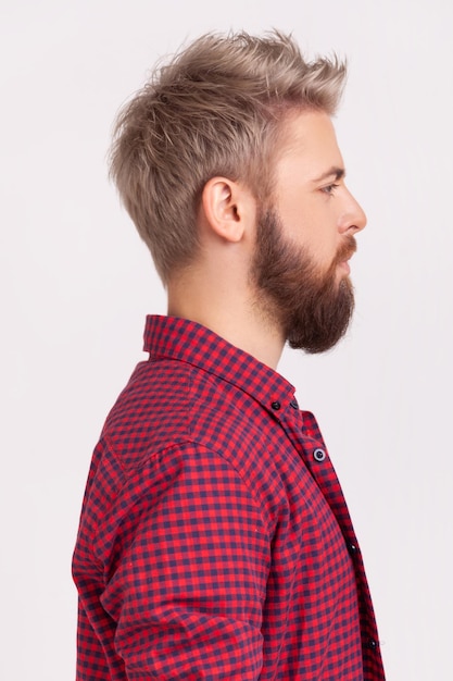 Profile portrait of confident bearded male with blond hair wearing red plaid shirt looking to side space with serious attentive face Indoor studio shot isolated on gray background