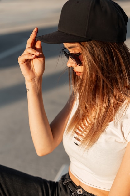 Profile portrait cool urban fashionable woman in dark sunglasses in white sexy top in stylish cap in sun in city. Beautiful urban girl posing in parking lot on sunny day.