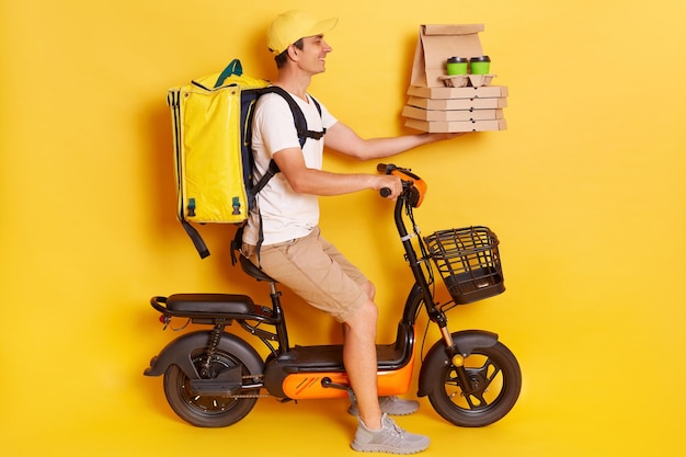 profile portrait of courier man wearing cap and tshirt driving electric bike motorbike handing pizza in cardboard and takeaway coffee isolated on yellow background
