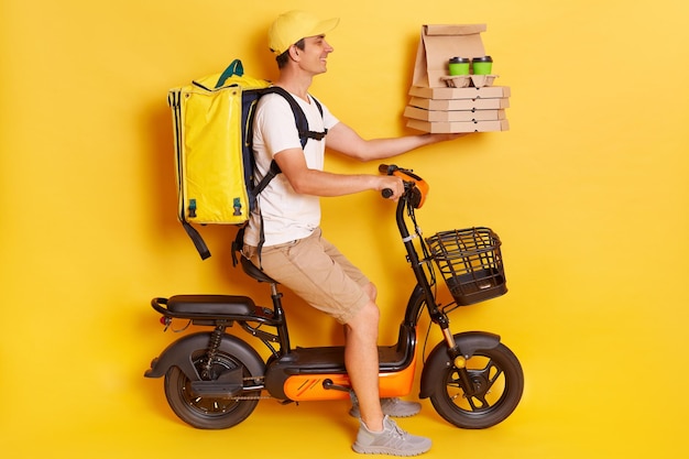 キャップとTシャツを着て電動自転車バイクを運転している宅配便の男のプロフィールの肖像画は、黄色の背景で隔離の段ボールと持ち帰りのコーヒーでピザを手渡します