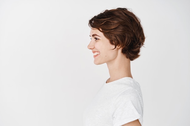 Photo profile portrait of healthy natural woman with short hairstyle, smiling and looking left at empty space, standing in t-shirt on white wall