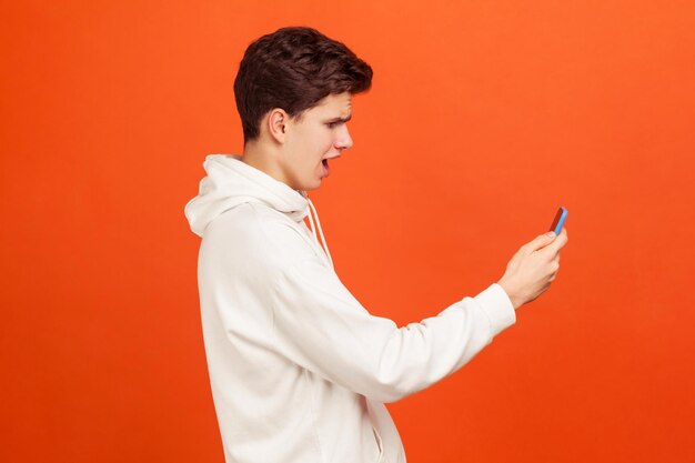 Profile portrait of shocked teenager in white casual sweatshirt looking at screen of mobile phone with excited face, receiving email with sale coupons. indoor studio shot isolated on orange background