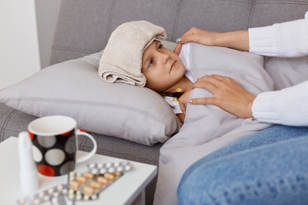 Profile portrait of sick little baby girl with influenza lying on pillow on sofa with wet towel on forehead, mother treating her child giving pills and tea, kid catching cold in wet weather.