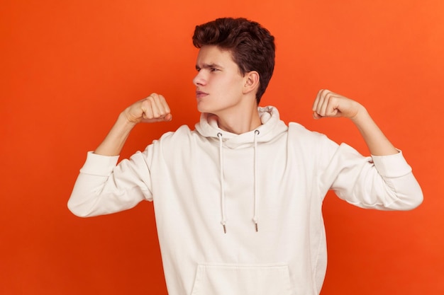 Profile portrait of teenager with stylish hairdo in fashionable hoodie demonstrating his arm muscles, strong and independent male person visiting gym. Indoor studio shot isolated on orange background