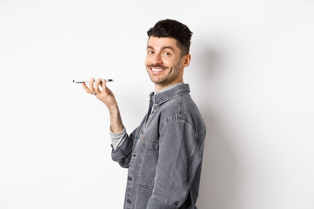 Ritratto di profilo di giovane uomo sorridente alla telecamera, registrazione vocale per tradurre la parola o lasciare un messaggio, parlando in vivavoce, in piedi su sfondo bianco.