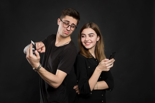 Profile portrait of young married couple, browsing information at their pdas, standing back to back, wearing casual outfits on black background.