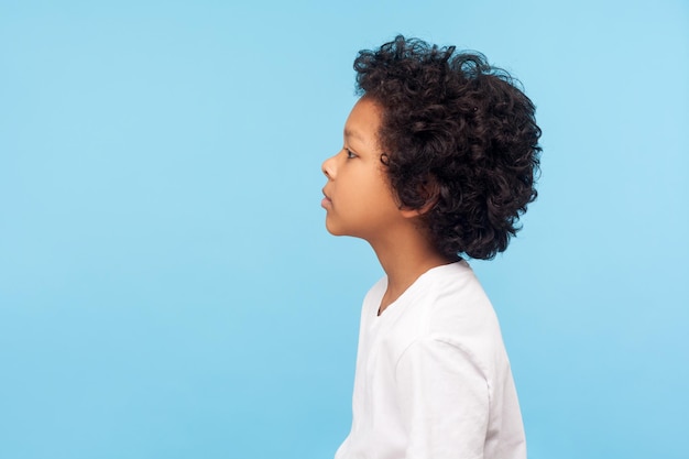 Profile of pretty nice little boy with stylish curly hair in Tshirt looking to side with serious attentive face calm pensive expression indoor studio shot isolated on blue background copy space