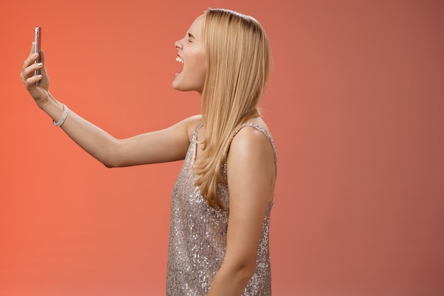 Profile shot funny carefree blond woman hold smartphone raised open mouth wide yelling recording video own shout scream, standing silver glamour dress near red background fool around.