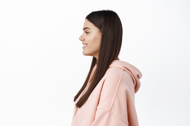 Profile shot of young brunette girl with fresh clean skin and long hair, looking left at copy space, standing against white wall