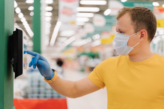 Profile shot of young man types on touch screen of self checkout wears protective rubber blue gloves poses against blurred supermarket background Consumerism shopping quarantine concept