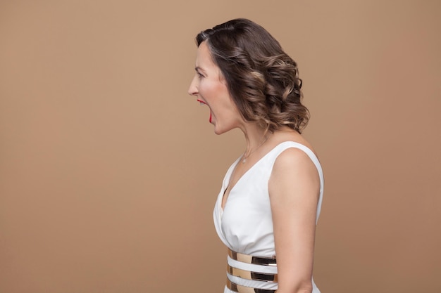 Profile side view of angry middle aged boss woman. Emotional expressing woman in white dress, red lips and dark curly hairstyle. Studio shot, indoor, isolated on beige or light brown background