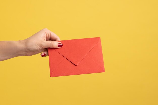 Profile side view closeup of woman hand holding red letter
envelope romantic message indoor studio shot isolated on yellow
background