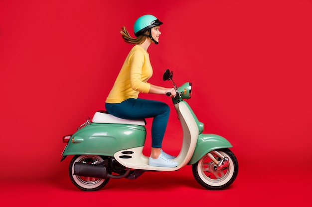 Profile side view of lovely cheerful girl driving moped on red background