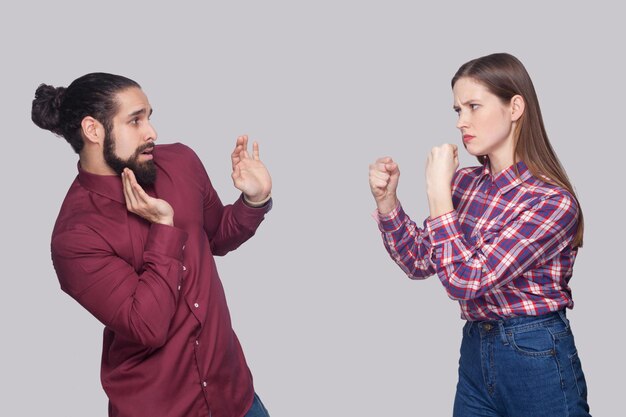 Photo profile side view portrait of angry woman standing with boxing fists and ready to attack at confused scared man with black collected hair. indoor studio shot, isolated on gray background.