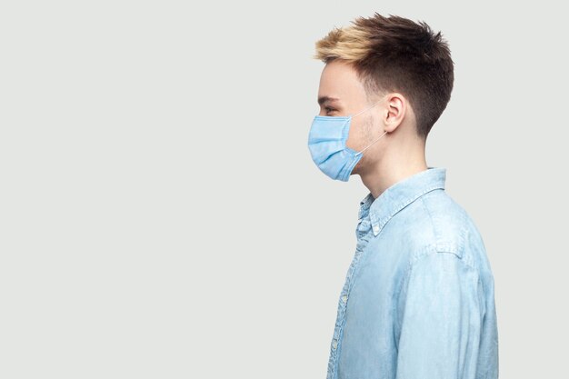 Profile side view portrait of handsome young man with surgical medical mask in light blue shirt standing and looking forward . indoor studio shot on grey background copy space.