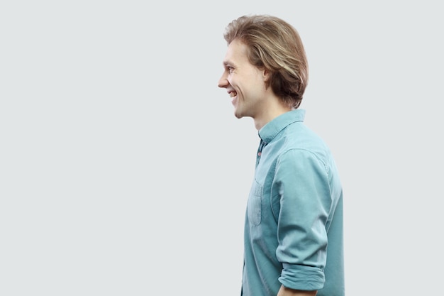 Photo profile side view portrait of happy handsome long haired blonde young man in blue casual shirt standing and looking with toothy smile. indoor studio shot, isolated on light grey background.