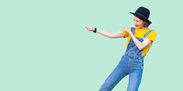 Profile side view portrait of pretty young hipster girl in blue denim overalls, yellow shirt and black hat standing with fist or pulling gesture. indoor studio shot isolated on light green background.