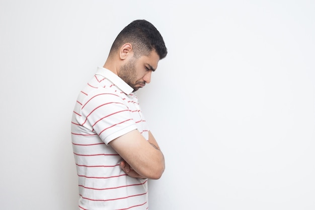 Profile side view portrait of sad alone o depressed bearded young man in striped t-shirt standing, holding his head down and feeling bad or thinking. indoor studio shot, isolated on white background.