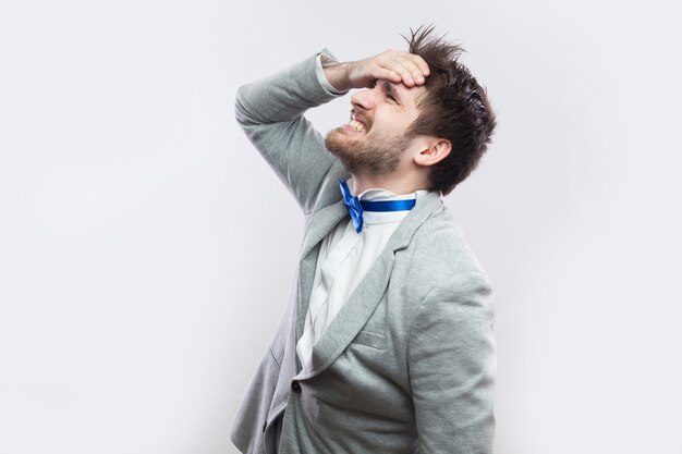 Photo profile side view portrait of sad handsome bearded man in casual grey suit and blue bow tie standing hopeless because he lose everything and mistaked. studio shot, isolated on light grey background.