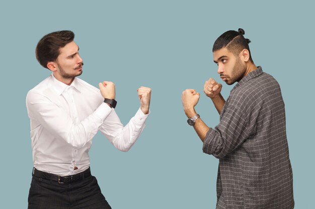 Profile side view of two angry boxer businessman, looking to each other with serious face and ready to attack. crisis and partnerships problem. indoor studio shot, isolated on light blue background.