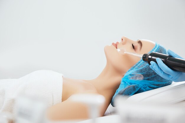 Profile of smiling woman in blue cap lying on couch in cosmetological clinic with closed eyes. Doctor's hands in blue gloves doing Darsonvalization procedure