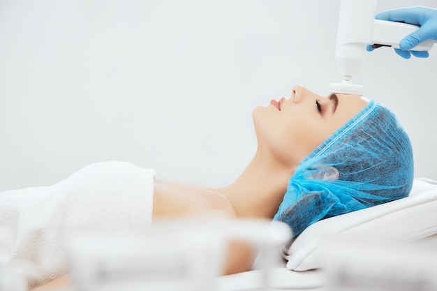 Profile of smiling woman in blue cap lying on couch in cosmetological clinic with closed eyes. Doctor's hands in blue gloves doing procedure of brossage