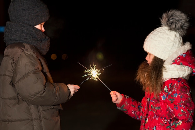 Profilo di due bambini piccoli svegli, ragazzo e ragazza in abbigliamento invernale caldo che tiene fuochi d'artificio sparkler brucianti nella notte oscura all'aperto copia sfondo spazio. anno nuovo e celebrazione del natale concetto.