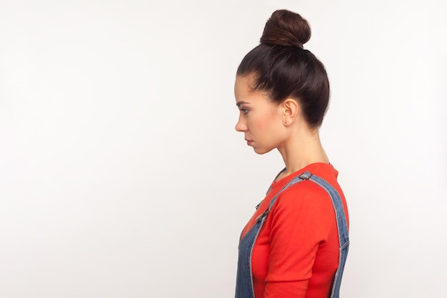 Photo profile of upset depressed girl with hair bun in denim overalls standing with head down and unhappy look, expressing sadness, worrying about problems. indoor studio shot isolated on white background