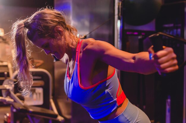 Profile view of caucasian woman in gym standing training biceps shoulder and back with pulleys