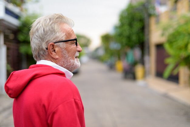 Foto vista di profilo di uomo barbuto senior bello felice che sorride mentre indossa gli occhiali all'aperto