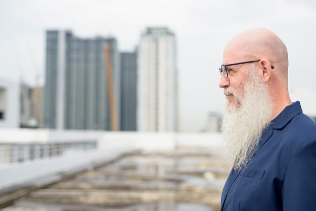 Вид в профиль зрелого лысого бородатого бизнесмена в очках на фоне города