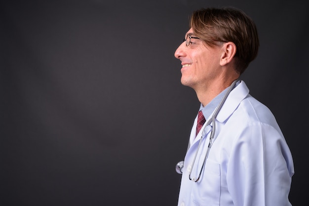 Profile view of mature handsome Italian man doctor against gray wall