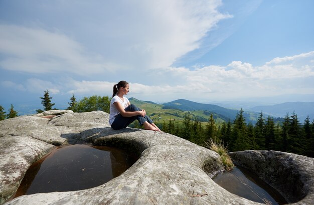 Profile view of tourist girl sitting on huge boulder with big puddle in the middle under beautiful cloudy sky