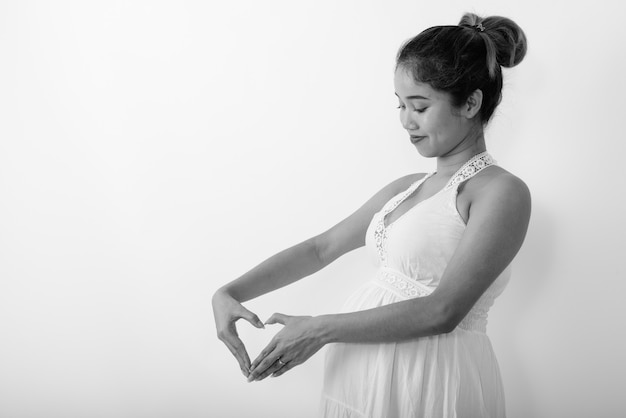 Vista di profilo della giovane donna incinta asiatica che fa il segno della mano del cuore accanto al suo stomaco contro il muro bianco