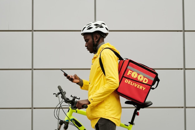 Photo profile view of young courier in helmet and uniform using smartphone